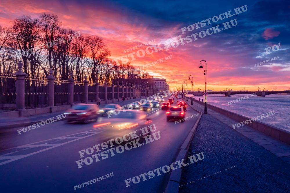 На фотографии изображена набережная Кутузова в Санкт-Петербурге на закате. Видна ограда Летнего сада, проезжающие автомобили и река Нева. Фотография подходит для использования в туристических материалах, статьях о Санкт-Петербурге и иллюстрациях о городских пейзажах.